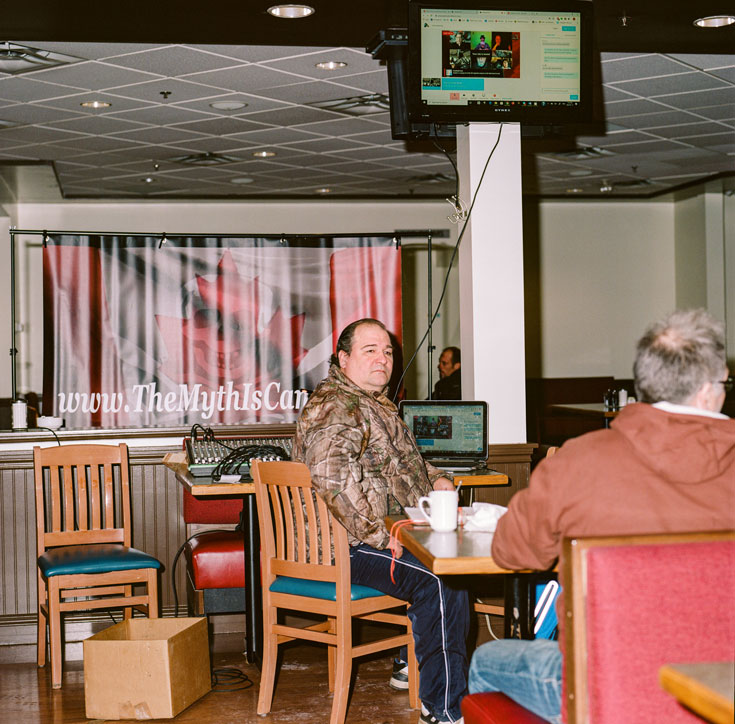An older man sits at a table in a restaurant, beneath a television.