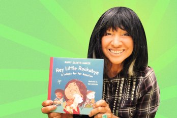A photograph of Buffy Sainte-Marie holding up her new picture book and smiling. The background is bright green with stripes radiating out from behind Buffy.