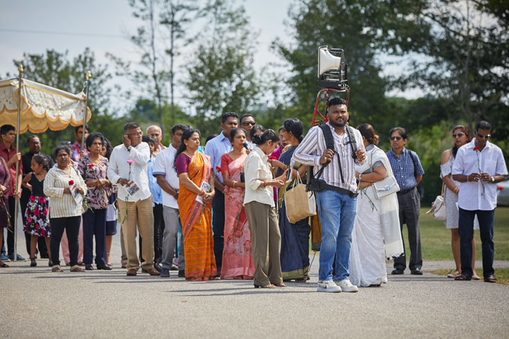 A procession of pilgrims stand outside. At the front of the group is a young man in a striped shirt, carrying a portable sound system on his back.