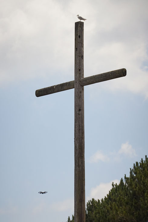 A large wooden cross against a blue sky. A seagull is perched on top of the cross. Nearby, a small drone flies through the air.